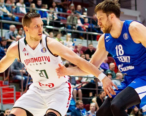 Два баскетболиста на площадке во время матча. Спортсмен в белой форме с надписью Локомотив-Кубань против игрока в синей форме с надписью Зенит. На заднем плане видны зрители на трибунах