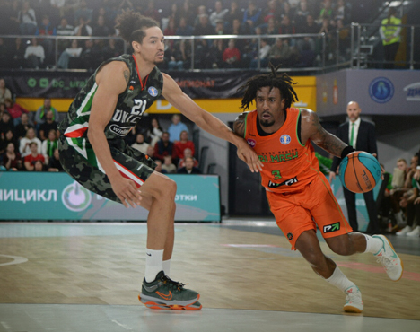На изображении два баскетболиста на игровой площадке. Игрок в зелёной форме защищается, а игрок в оранжевой форме, держа мяч в правой руке, пытается его обыграть. На заднем плане видны зрители