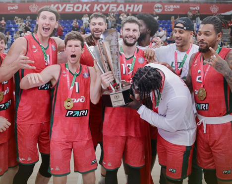 На изображении группа баскетболистов в красной форме празднует победу. Они держат кубок и носят медали. Эмоции радости и торжества. На заднем плане видны зрители и баннеры