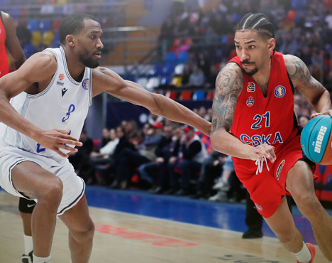 На изображении запечатлен баскетбольный момент: игрок в красной форме идет в нападение с мячом, защищающийся соперник в белой форме пытается его остановить. На заднем плане видны зрители трибун